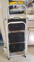 Two folding step stools stacked, showing white metal frames, black textured steps, and carrying handles at top.
