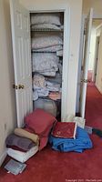 Photo showing open closet shelves stacked with folded comforters, pillows, and various linens with additional folded linens on floor in front of closet.