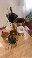 Full view of the lot showing the umbrella stand with umbrellas, brass planters, wastebasket, and live plant on hardwood floor near window.