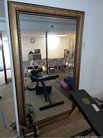 Full view of large wall mirror with gold ornamental frame reflecting a basement exercise area and dining table.