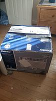 Boxed Danby window air conditioner placed on wooden floor next to a dresser and door.