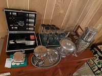 Overview of the barware collection on wooden sideboard including glasses, ice buckets, metal tray with containers, and cocktail shaker set in case.