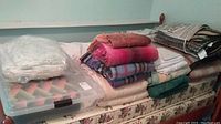 Stack of folded bedding and blankets on a mattress, some blankets are brightly colored while others are neutral tones.