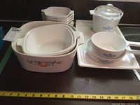 Photo showing large square baking dish with handles, three rectangular baking dishes nested inside it, four small square dishes stacked, covered casserole, serving bowl and tray arranged on table.