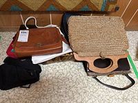 Photo shows a pile of ladies handbags and a pair of black gloves on a floor with a patterned carpet in the background.