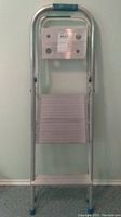 Front view of closed aluminum step ladder showing one wide step and top handle with blue grip.