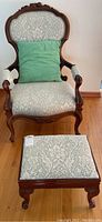Full view of the vintage upholstered chair and its matching foot stool against a light wall and wooden floor. Chair has carved wood framing and light damask fabric upholstery with a green pillow.