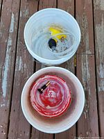 Top-down photo of two white 12L buckets on wooden floor. One bucket contains siphon pump with yellow handle and clear tubing, the other bucket holds a red hose nozzle and black connector.
