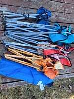 Photo of folded camping chairs and folding stools laid on a wooden deck, showing metal frames and fabric seats in various colors including blue, red, orange, and black.