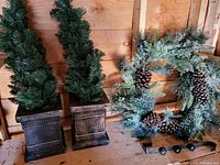 Two artificial lighted pine trees in decorative planters beside an artificial Christmas wreath with pine cones and snowy flocking on a wooden surface.