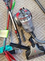 Photo showing Hoover vacuum with dustbin, hose, two black vacuum attachments, red broom with bristle head, green Swiffer mop and green hand brush on carpet.