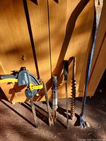 Five metal tools including two crowbars, a two-prong tool, a large auger bit, and a clamp, placed against wooden cabinet.
