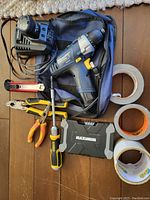 Items laid out on floor including cordless drill, batteries, charger, pliers, screwdriver, utility knife, and three rolls of tape next to blue carrying bag.
