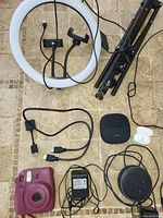 Overhead photo of ring light, tripod, charging cables, Bose speaker, purple instant camera, and other small electronics on tiled floor.