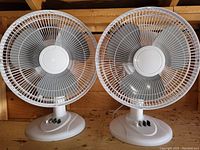 Two identical small white tabletop fans with plastic blades and wire cage front guards, placed side by side on a wood surface.
