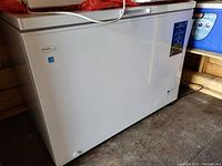 White Danby chest freezer with closed lid, placed on a wooden floor next to a blue cooler and wooden crates.