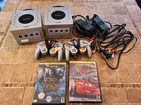 Two silver Nintendo GameCube consoles, power adapters, AV cables, controllers, and game cases laid out on tiled surface