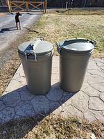 Two gray plastic garbage bins with round lids and latch closures. Positioned outdoors on stone pavement with grass and fencing background. One lid missing a latch, visible bite marks on one bin and lid.