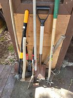 Eight gardening tools leaning against wooden wall, showing different types and sizes including pruner, shovels, hoe, and cultivator.