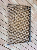 One of the two Evenflo expandable baby gates with brown criss-cross lattice and black frame, hooks visible at top and bottom, laid on wooden floor.