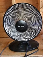 Black Noma electric heater with protective grill and adjustable dial on wooden floor background.