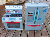 Photo shows two Little Tikes toy kitchen appliances placed on a wooden surface. On the left is a stove featuring control knobs, burners, a grill area, and an oven door with a clear window and red handle. On the right is a refrigerator with double doors, blue handles, and ice and water dispenser decals.