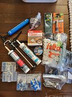 Overview of caulking gun, multiple tubes of glue and caulking, assortment of screws and plugs, and other small hardware parts laid out on wooden floor.