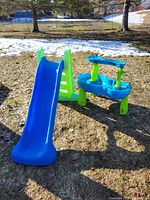 Blue and green plastic slide alongside a blue and green water play table set outdoors on grass with some snow patches visible in background.