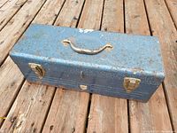Closed vintage blue metal toolbox placed on wooden deck showing rust and chipping paint.