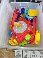 Large clear plastic storage tote filled with colorful VTech Smart Wheels track components including a red roundabout with road patterns and various connected track pieces