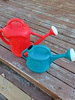 Two large plastic watering cans, one red and one teal, placed on a wooden deck with floral designs and detachable sprinkler spouts.