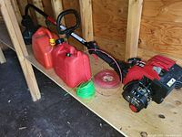Photo showing a gas-powered whipper snipper with handle and curved shaft, two red gas cans, and two coils of trimmer line on a wooden shelf inside a shed.
