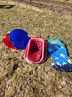 Four winter sled-related items placed on patchy grass: two round snow saucers (one mostly blue with red interior, one red), a red and black checkered inflatable sling-type sled, and a fabric sled with a blue top and polar bear print.