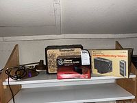 Four vintage items arranged on a shelf; a vintage Magnajector in original box, a GE Zonar burglar alarm box, and another infrared photorelay/alarm box.