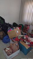 Photo showing stacked boxes, plastic bins, and bags with assorted Christmas decorations, gift bags, and linens next to window with curtain.