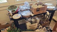 Photo of several wall clocks, glassware, plates, and boxes with smaller items on and under a table.