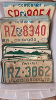 Photo showing multiple vintage Colorado license plates featuring Centennial 76 and Colorful 73 designs, stacked in plastic container.