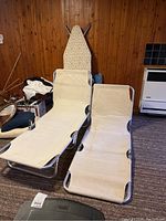 Photo of two beige folding cots with metal frames, camp chair, and ironing board visible in background against wooden wall.