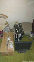 Photo showing boxed Texas Instruments Home Computer, beige Brother electric typewriter, and Sony Trinitron monitor placed on green carpet alongside a brown box and some other items