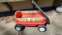 Red Fisher Price toy wagon with black handle and wooden side rails viewed from side outdoors on pavement.