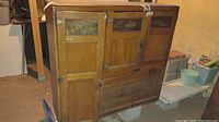 Front and side view of top portion of wooden Hoosier cabinet showing three doors with glass panels and wooden panels below, plus tambour roll door at base. Visible water damage and signs of wear.