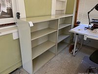Two matching light gray metal bookcases with several shelves each, positioned side-by-side against a wall near a desk.