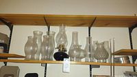 Shelf display with multiple glass chimneys, one dark blue base oil lamp with chimney, and another glass oil lamp base without chimney, plus additional chimneys and lamp parts.