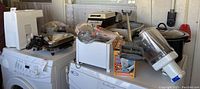 Wide view of assorted small kitchen appliances on washing machine and dryer including waffle iron, toaster, utensil sharpener, crockpot, curly cutter maker, and electric skillet.