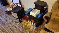 Wide shot of all items including Sterilite 2-drawer unit, several large black storage totes, black rubbish bin, and metal wire racks on carpeted floor.