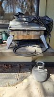 Front angled view of BHG stainless steel tabletop gas grill with standard propane tank on ground beneath it and two small propane tanks to the side.