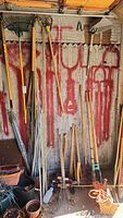 Full view of yard tools leaning against a wall, including rakes, pickaxe, axes, post hole diggers, weeders, and handles of various tools.