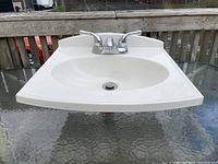 Front view of a white wall-mounted bathroom sink with chrome dual-handle faucet on a glass table outdoors.