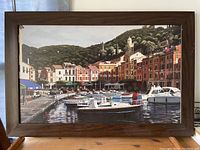 Framed painting showing a harbor scene with boats docked and colorful buildings with a green hill background, in a brown frame.