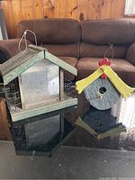 Two wooden garden items: a large bird feeder with green roof and glass sides on the left, and a small colorful bird house with yellow roof and red comb on the right